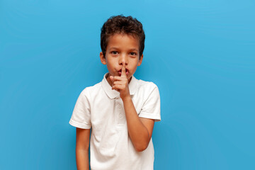 African-American boy in white polo asks to be quiet and holds his finger near his lips on blue isolated background