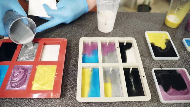 Craftsman pouring silver colour epoxy resin from plastic cup into silicone mold and stirring with wooden stick, process of creation of different colours samples for furniture decoration or accessories
