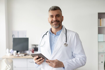 Professional help. Happy middle aged male general practitioner standing with tablet and smiling at...