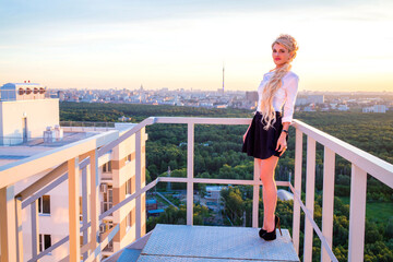 Girl with beautiful long blond hair in white blouse and short skirt is standing on fire escape on...