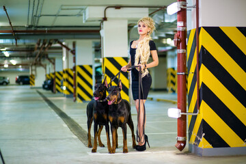 Beautiful girl with long blond hair in dress with two Doberman on leashes in the underground parking