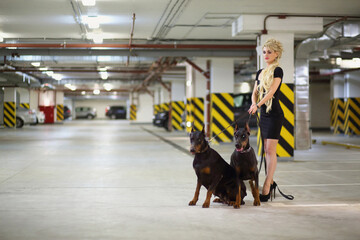 Beautiful girl with long blond hair in black dress holding two dogs on leash in the underground...