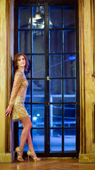 Woman in short shiny dress with sequins leaning on wall near the glass door in an ancient palace interior