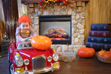 A little boy in an orange cap in red car carries big pumpkin on the hood of car in front of...