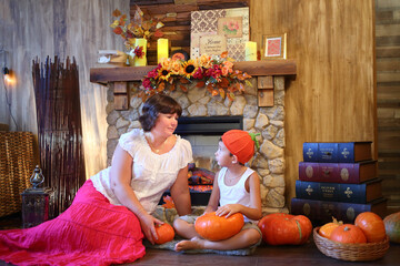 Mother and son in orange cap with pumpkins are sitting in front of fireplace