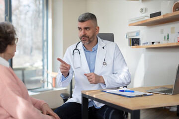 Fototapeta premium Middle aged male doctor consulting senior female patient in clinic, GP have consultation with elderly woman client in private hospital