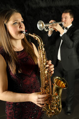 Woman playing on saxophone in front of man with trumpet in black room