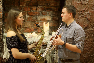Two musicians with wind instruments in their hands look at each other in front brick wall with...