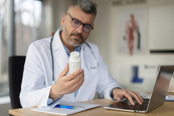 Obraz premium Middle aged man doctor holding jar of pills in hand and using laptop, surfing internet and writing prescription for patient, sitting at desk in clinic