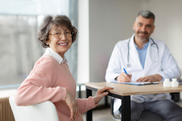 Positive senior elderly woman turning back and smiling while her middle aged male practitioner sitting on the background