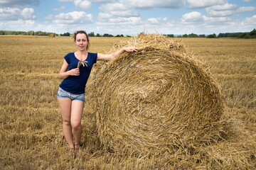 Naklejka na ściany i meble Young woman in shorts and t-shirt is standing near huge haystack.