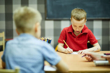 Children draw with pencils sitting at desk in classroom, focus on boy in red shirt.