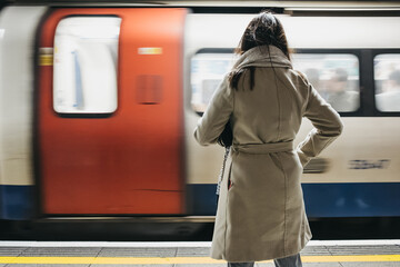 Rear view of a woman standing on a London Underground station platform, moving train on the background.