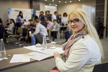 Young blond woman sits at table with papers in auditorium, shallow dof.