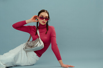 Fashionable confident woman wearing pink wraparound sunglasses, turtleneck, white skirt, with trendy metallic leather bag posing on blue background. Studio fashion portrait. Copy, empty space for text