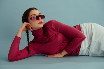 Fashionable confident woman wearing trendy pink wraparound sunglasses, turtleneck, white skirt, posing on blue background. Studio fashion portrait. Copy, empty, blank space for text