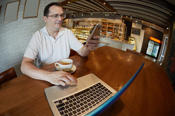 Man works at table with laptop, smartphone and cup of tea in cafe.