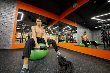 Man sits on big inflatable finess ball holding dumbbells in hands in gym.