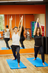 Man and woman stand on mats in yoga positions in gym.