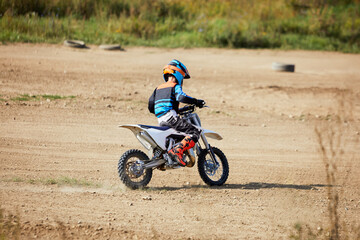 Boy in suit and helmet rides sports motorbike on dusty dirt road.