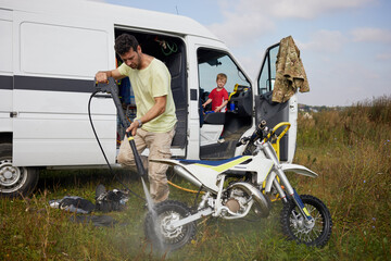 Man washes motorbike after training outdoor near minibus, little son looks at him from driver cabin.