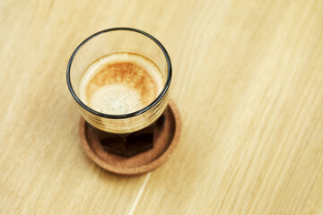 Black coffee with foam in glass on saucer on wooden desk.