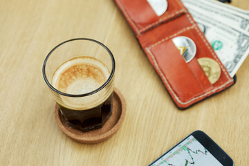Glass of coffee, smartphone with financial charts and red wallet with coins and banknotes on wooden desk, focus on glass edge.