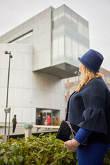 Blonde woman in blue suit and hat stands looking at silvery building.