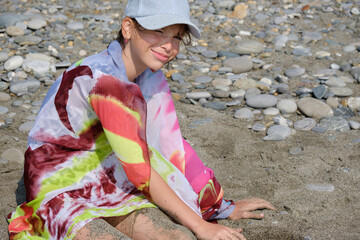 A teenage girl, wearing a cap and pareo, sits on a pebbled beach and looks into the camera.