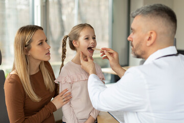 Angina, tonsillitis, cold and flu. Man pediatrician checking patient little girl throat, family mother and daughter attending clinic