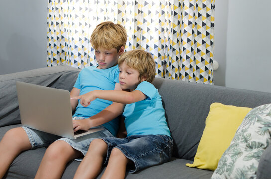 Horizontal photography of blond cute brothers in blue t-shirts sitting on the couch looking and pointing at the screen of a laptop watching videos or a movie. Children and technology concept.