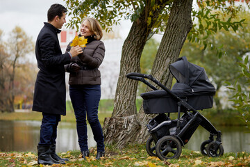 Smiling man and woman with perambulator stand in autumn park on river bank.