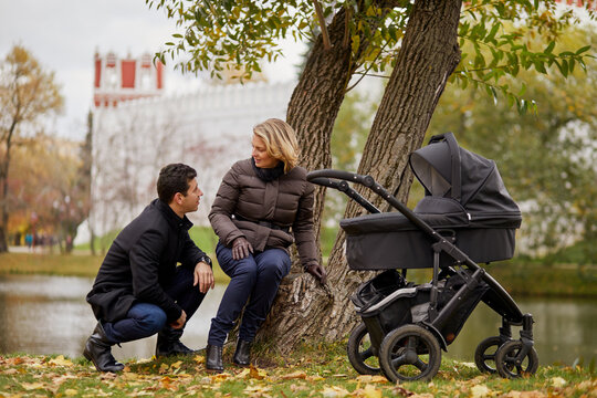 Man and woman rest near river during walk with perambulator in autumn park.