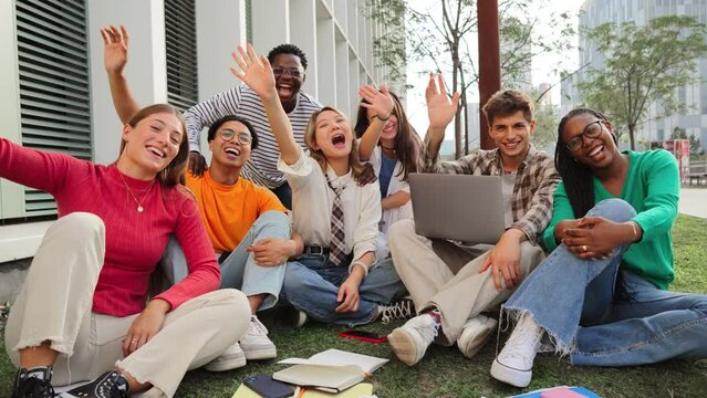Big group of real happy teenage students sitting at university campus lawn, greeting and looking at camera together. Positive multiracial teenagers laughing, talking, gesturing and waving hands.