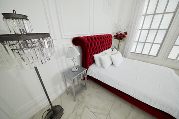 White bedroom interior with large red bed, flowers, torchiere and window.
