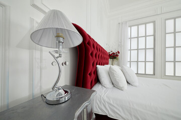 Lamp on bed-table and large red bed in white bedroom with latticed window.