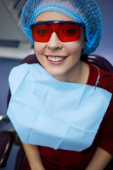 Smiiling woman in protective red glasses sits on dentist chair in office.