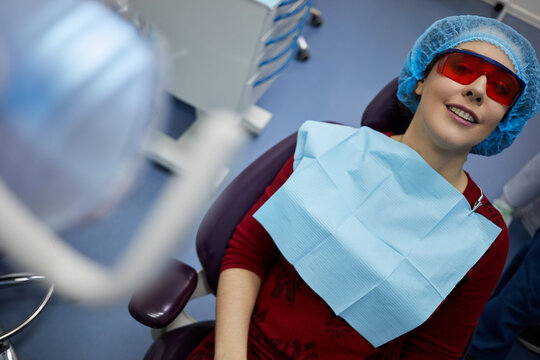 Smiiling woman in protective red glasses sits on stomatologist chair in office.