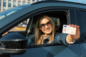 smiling young woman showing her new driver license out of car window after successful test at driving school - 762304852