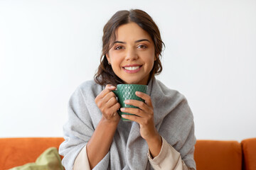 Beautiful woman wrapped in blanket holding tea cup and smiling at camera