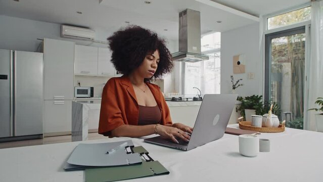Wide shot of Latin female freelancer working on laptop sitting at table in studio flat