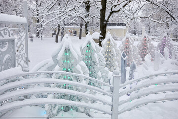 Artificial fir trees covered with snow and illuminated by garlands in winter park.