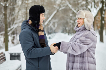 Smiling man and woman stand holding hands and looking at each other in winter park.