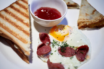 Fried pieces of sausage and egg, toasts, pancakes and jam in bowl on white dish.