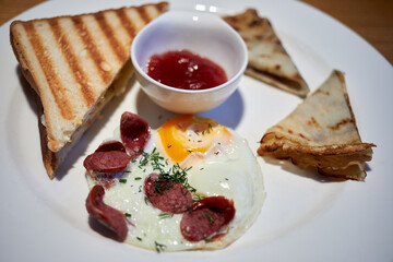 Fried egg with pieces of sausage, toasts, pancakes and jam in bowl on white dish.