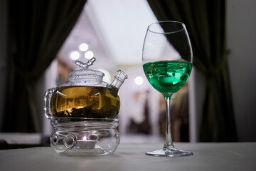 Transparent glass teapot on candle heater and wineglass with green beverage with ice on table.