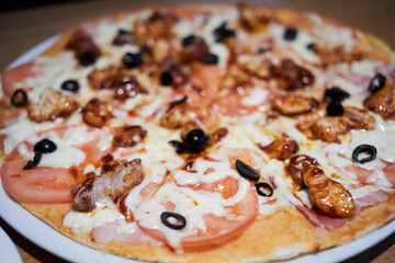 Close-up pizza with ham, tomatoes, cheese, black sliced olives on white plate.