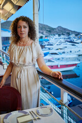 Smiling curly woman stands leaning on chair back and railing in floating cafe with bay view.