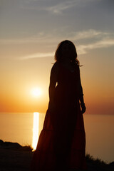 Silhouette of woman in dress standing at seashore on sunset.
