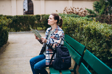 Fototapeta premium Beautiful stylish 30s woman in trendy wear spending time on street using paper map. Happy girl sitting on the bench. Urban lifestyle concept, travel banner